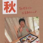 5品のおかずの器が置かれた四角のお盆を前にピースサインをする母の画像